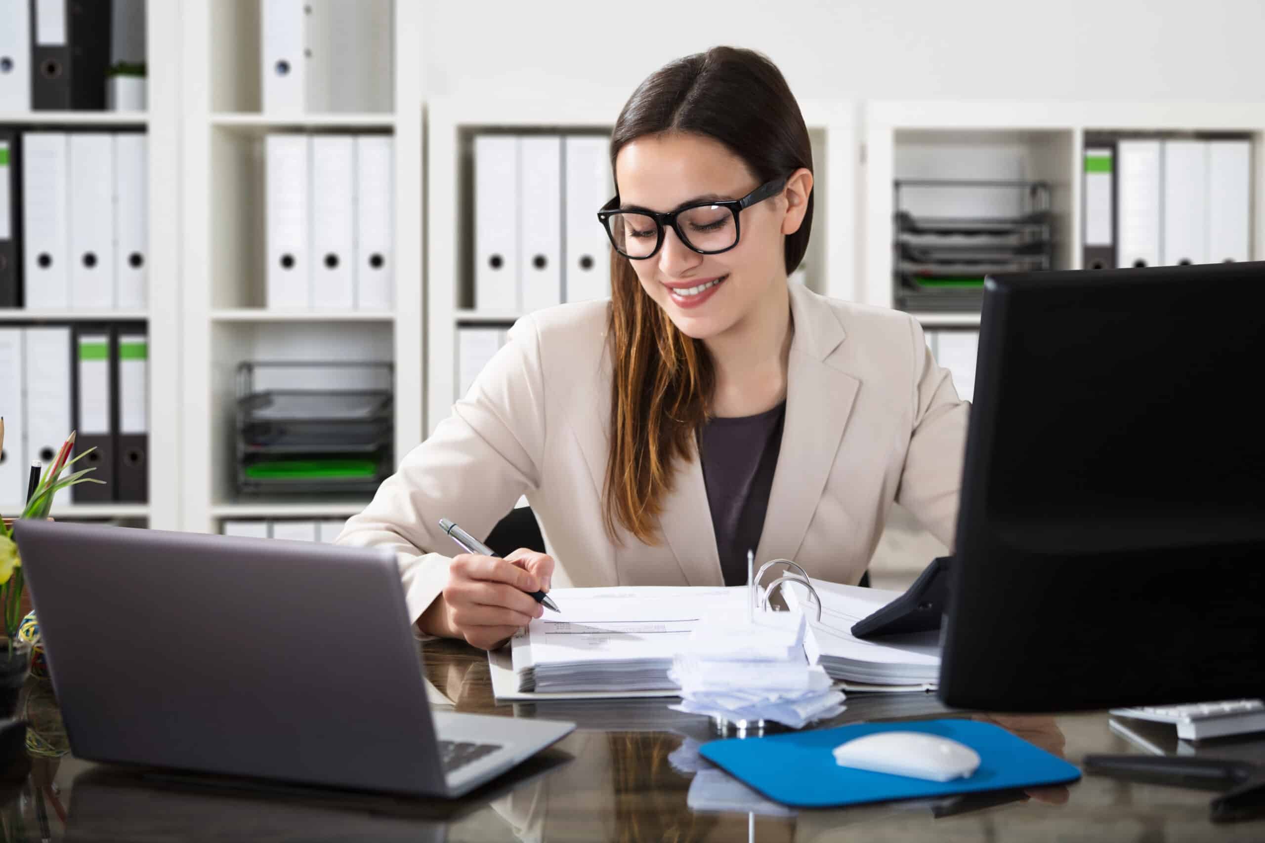 accountant working on a stack of papers at her desk sitting behind two computers 