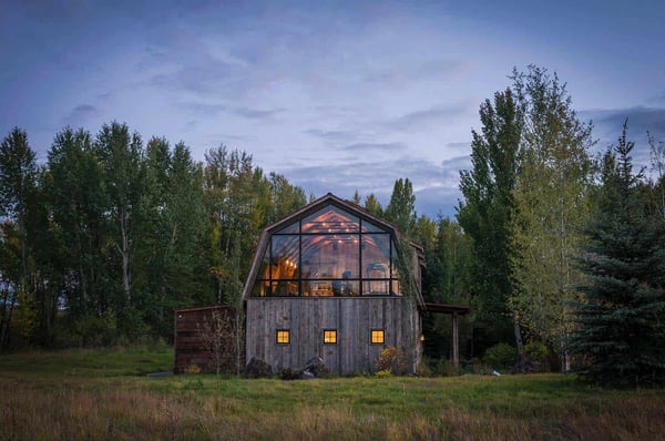 A barn style guest house with large glass window and verticle siding surrounded by green trees and a blue sky.