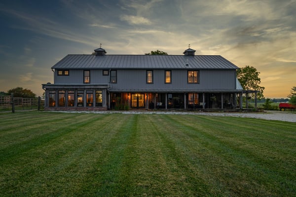A black modern barndominium with a big grass yard in front with a sunset sky in the background.