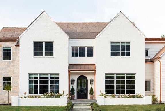 A white brick home with two symmetrical gables, large gridded windows, and a central entryway with a black arched door.
