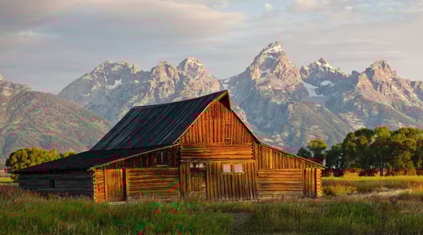 Old barn house illuminated by a rising sun off camera sitting in the middle of a field in front of a mountain range.