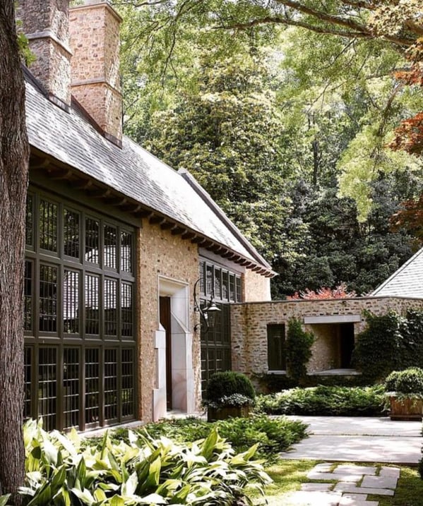 A transitional style stone home with modern black grid windows surrounded by green trees and plants