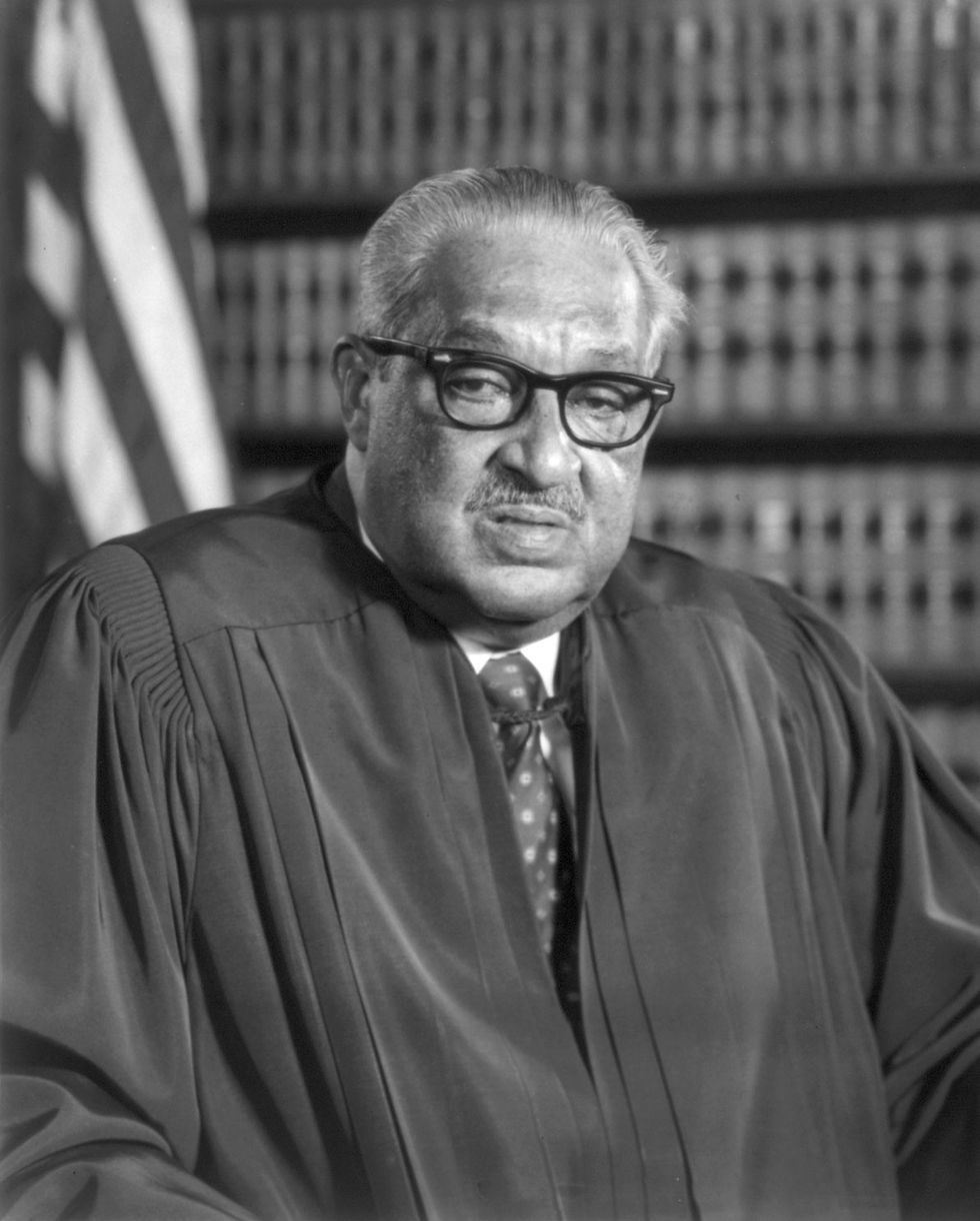 This is a black and white portrait of Thurgood Marshall, a Black man with short, gray hair. He's wearing judge's robes and glasses, and he's standing in front of a bookcase and an American flag.