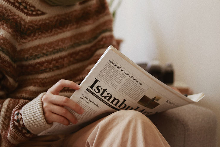 This is a picture of a woman reading a newspaper. She's sitting in a chair, she's wearing a brown striped sweater and tan pants, and she's reading a newspaper.