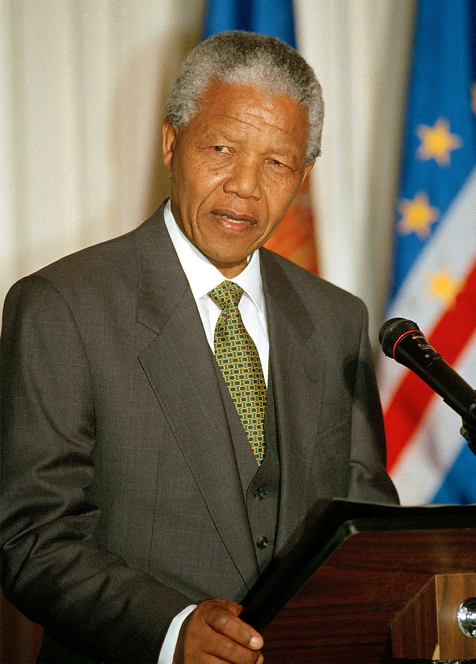 This is a portrait of Nelson Mandela, a Black man. He has salt and pepper hair, and he's wearing a gray suit, a white dress shirt, and a green tie. He's standing behind a podium, and there are a couple of large flags behind him.