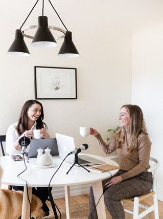 This is a picture of two women podcasting. They're both sitting at a table, in front of computers and microphones.