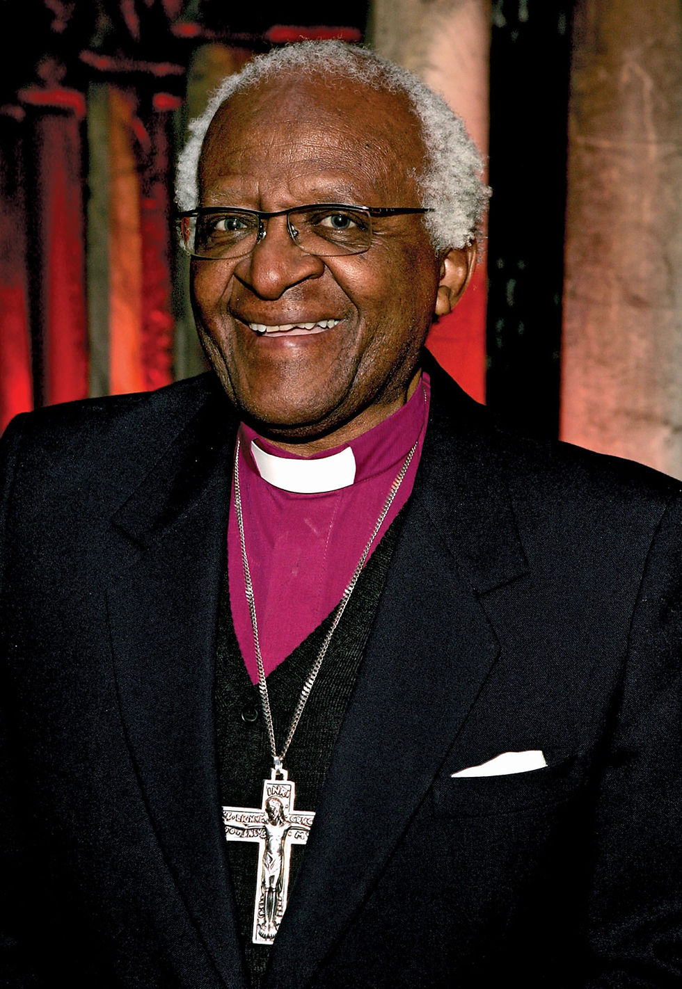 This is a portrait of Desmond Tutu, a Black man. He's pictured wearing a magenta shirt with a white Clerical collar, a black suit jacket, and a large silver crucifix around his neck. He also wears glasses and has short, silver hair. 
