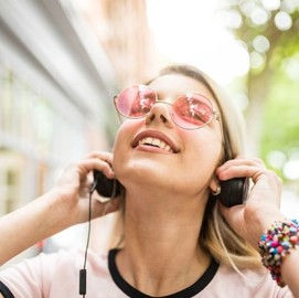 This is a picture of a blonde-haired woman walking outside. She's wearing rose-tinted, round glasses and headphones. 