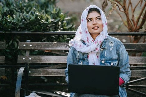 This is a picture of a woman sitting on a bench with a laptop on her lap. She has dark hair and a scarf wrapped around her head, and she's wearing a denim jacket. 