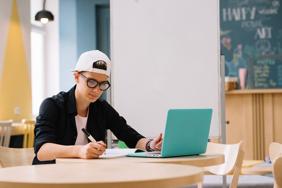 This is a picture of a male student sitting at a table in front of an open laptop. He's writing something in a notebook. 