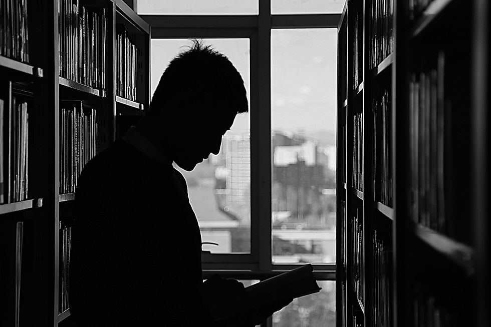 This is a black-and-white picture of a man standing in front of a bookshelf, reading a book. 