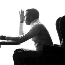 This is photograph of Chinua Achebe, a Black man. The picture captures his profile as he sits at a desk. He is wearing a light colored turtleneck and dark-colored pants. 