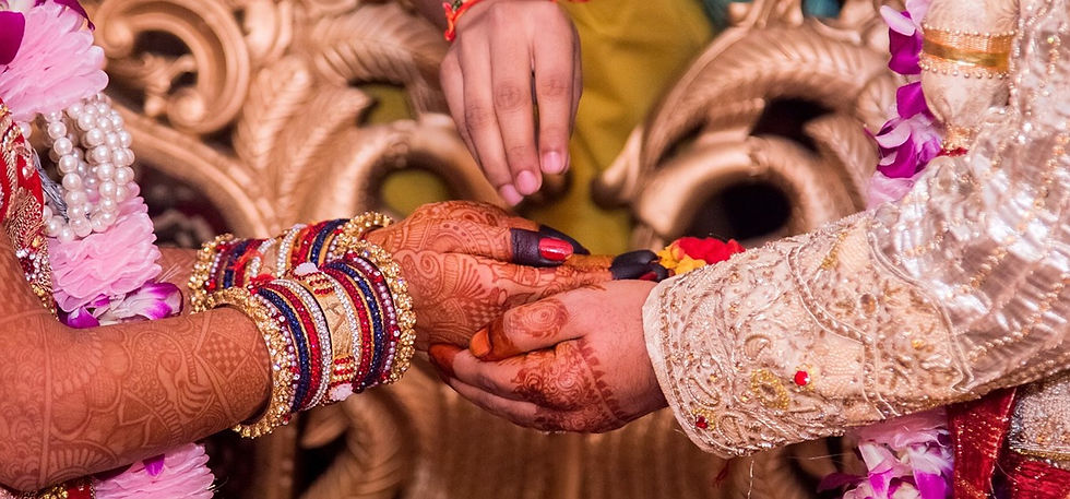 This is a picture of a man and a woman who are both holding hands. The woman's hands are covered in brown Henna tattoos, and she's wearing several bangles on both of her wrists. The man is also wearing some Henna tattoos, but only over his knuckles. Both are wearing ornate clothes, but no one's face is pictured. Above the hands is another hand wearing pink nail polish. The fingers are pinched together, as if the person is sprinkling something over the hands. 