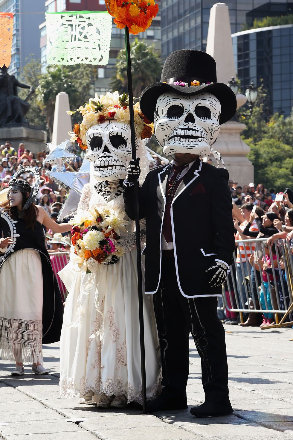 This is a picture of a man and a woman wearing costumes for a Day of the Dead celebration. Each of them is wearing an oversized fake skull over their heads. The man is wearing a black suit and top hat, and the woman is wearing a white dress and a wreath of flowers on her head. She's also carrying a bouquet of flowers in her hands.
