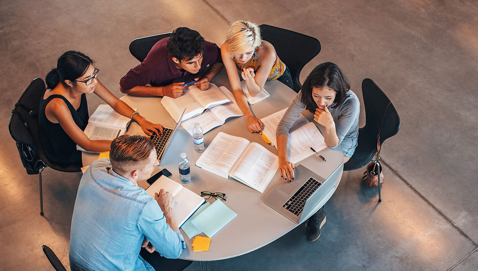 This is a picture of five students sitting at a table studying together. The table is covered with books, laptops, pens, water bottles, and a cell phone.