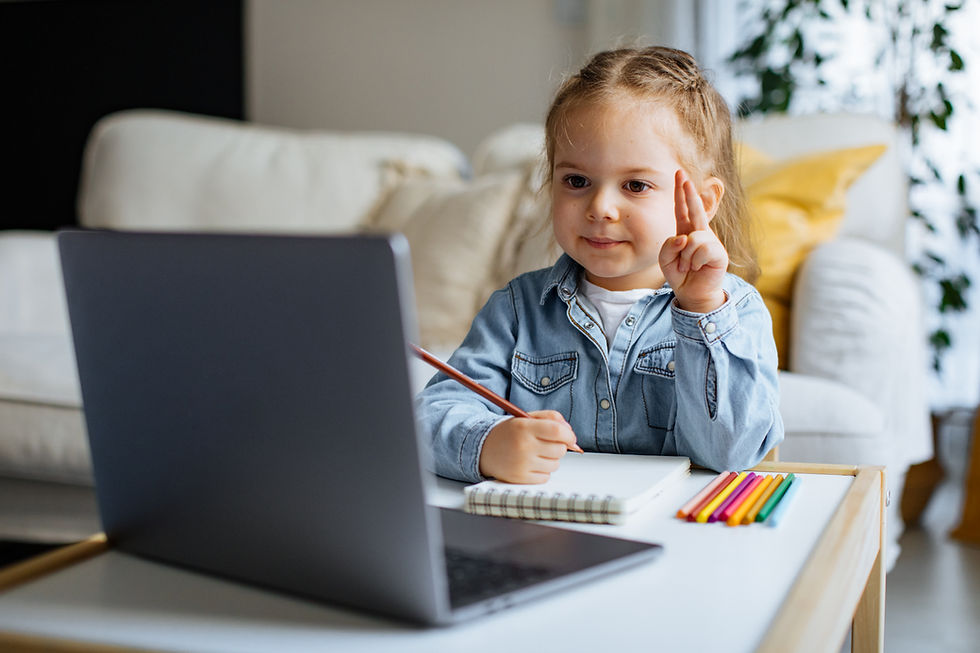 This is a picture of a young girl sitting at a table in front of a computer. She's writing on a notebook with a colored pencil.