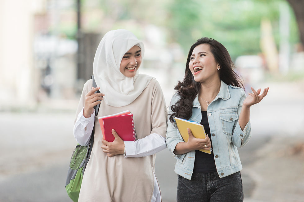 This is a picture of two students walking together and laughing. The woman on the left is wearing a white hijab and a long-sleeved, cream-colored dress. She's also holding a green backpack and a red book. The woman on the right is wearing a dark-colored dress and a light-colored denim jacket. She's holding a yellow notebook.