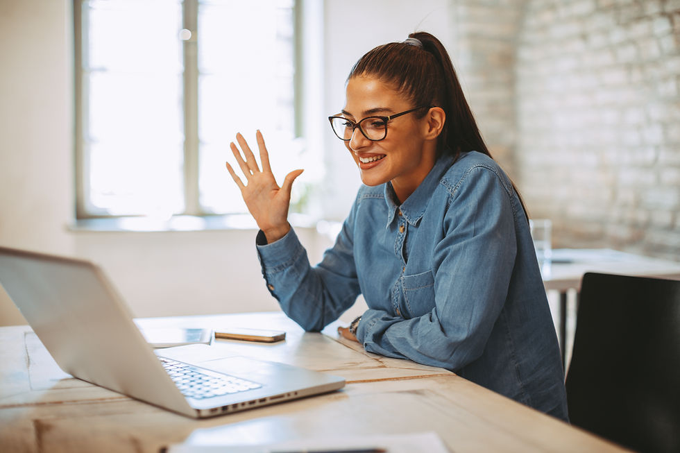 This is a picture of a woman sitting at a table, in front of an open computer. She's waving to the person on the other side of the screen. 