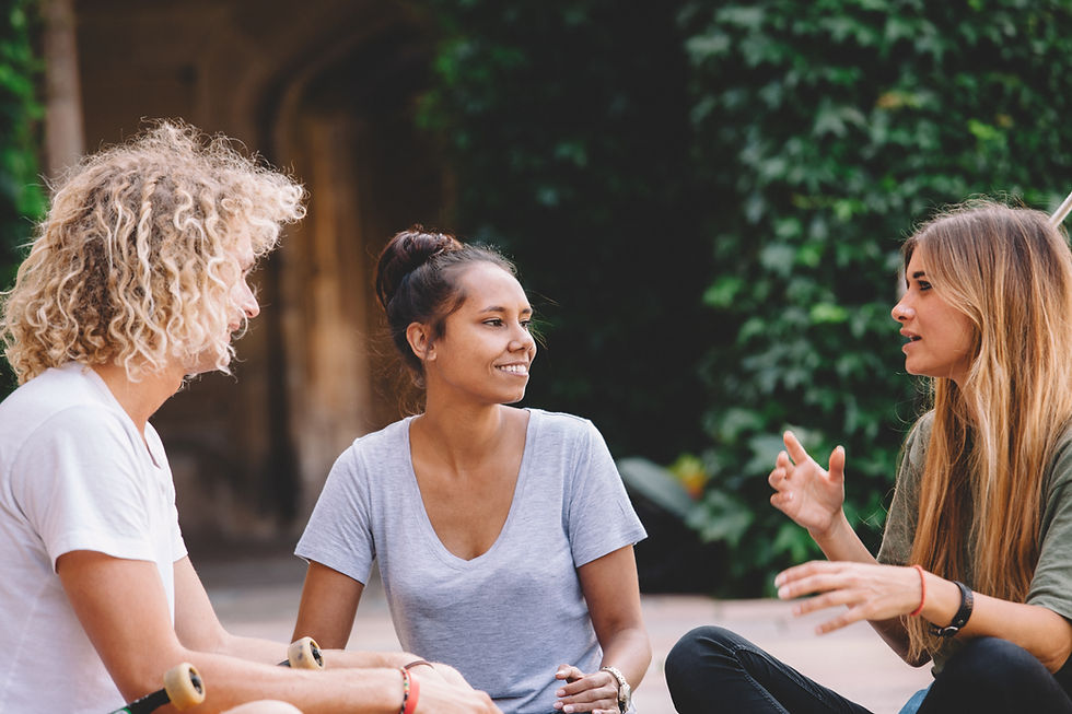 This is a picture of three women sitting together outside in a circle. One woman has curly blond hair and light skin, and she's wearing a white t-shirt. The second woman has her dark brown hair done in a bun. She has tan skin, and she's wearing a pale lavender t-shirt. The last woman has brown hair with blond highlights. Her skin is tan, and she's wearing an olive green shirt. In the background, there is a wooden wall with dark green ivy growing over it.