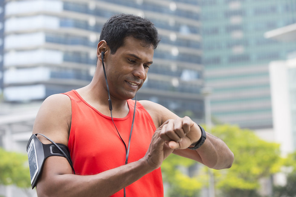 This is a picture of a man who is exercising. He's wearing an orange tank top, earbuds, an arm band with a cell phone, and a fitness tracker. He's looking at his fitness tracker. 