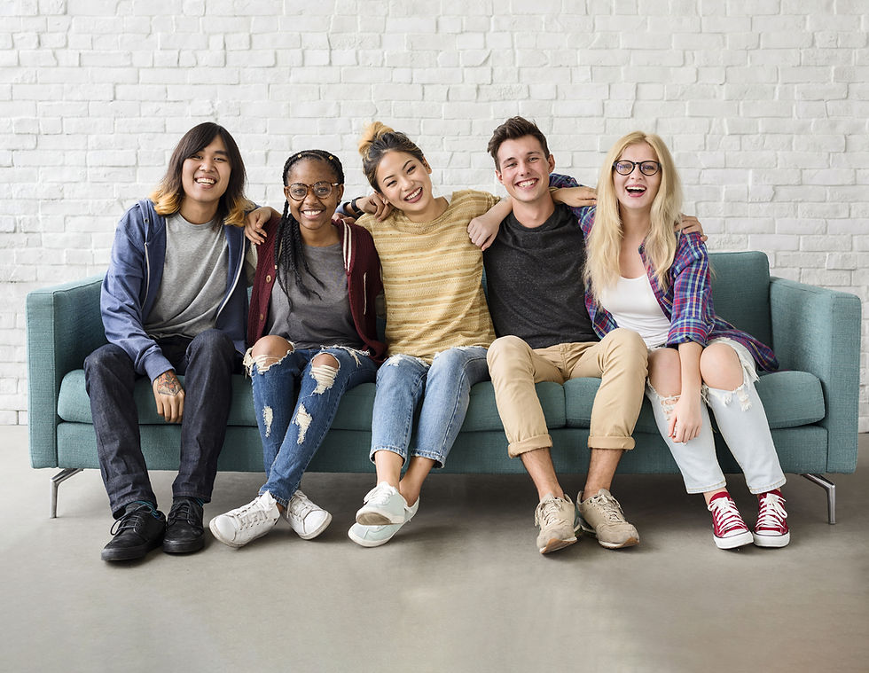 Five friends are sitting on a light green couch against a white brick background. Everyone is wearing jeans, t-shirts, jackets, and sneakers. From left to right, there is a Hispanic man, a Black woman, and Asian woman, a White man, and a White woman.