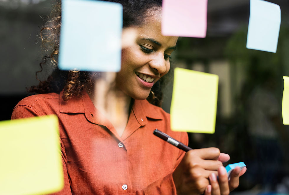 This is a picture of a Black woman writing something on a sticky note. She's wearing a dull orange top, and her dark, coily hair is pulled back. In between her and the camera is a pane of glass that has three yellow sticky notes, two blue sticky notes, and a pink sticky note stuck to it. 