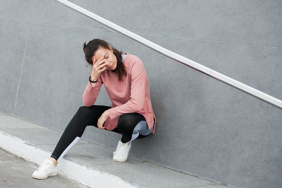 This is a picture of a young woman sitting against a gray wall. She has thin, straight brown hair tied into a ponytail. She's also wearing a light pink long-sleeved workout top, black exercise pants, and white tennis shoes. 