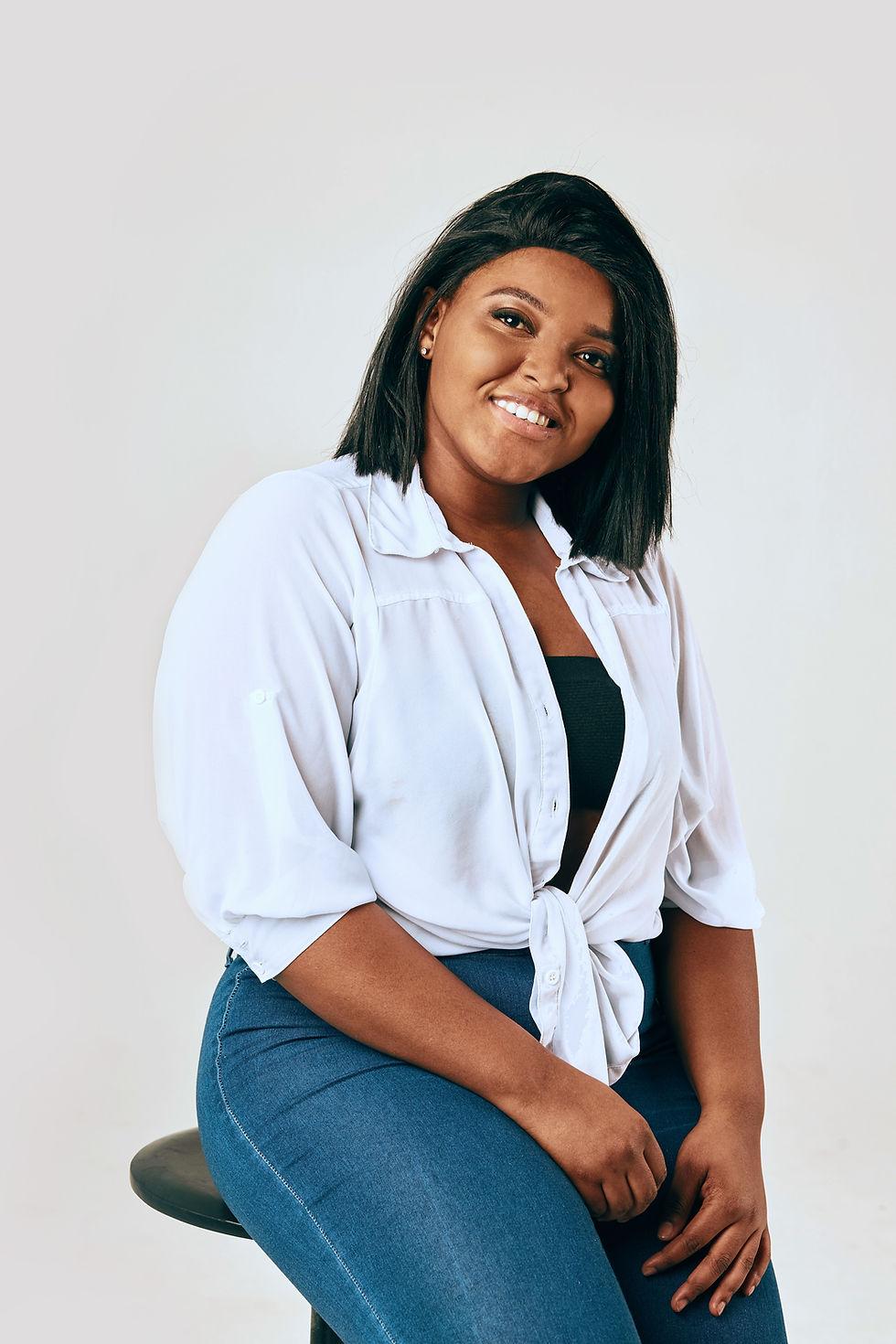 This is a picture of an African-American woman sitting on a black stool against a white background. She has short, straight, thick black hair. She's wearing a blue shirt with a white top over it, and she's earing blue jeans. 