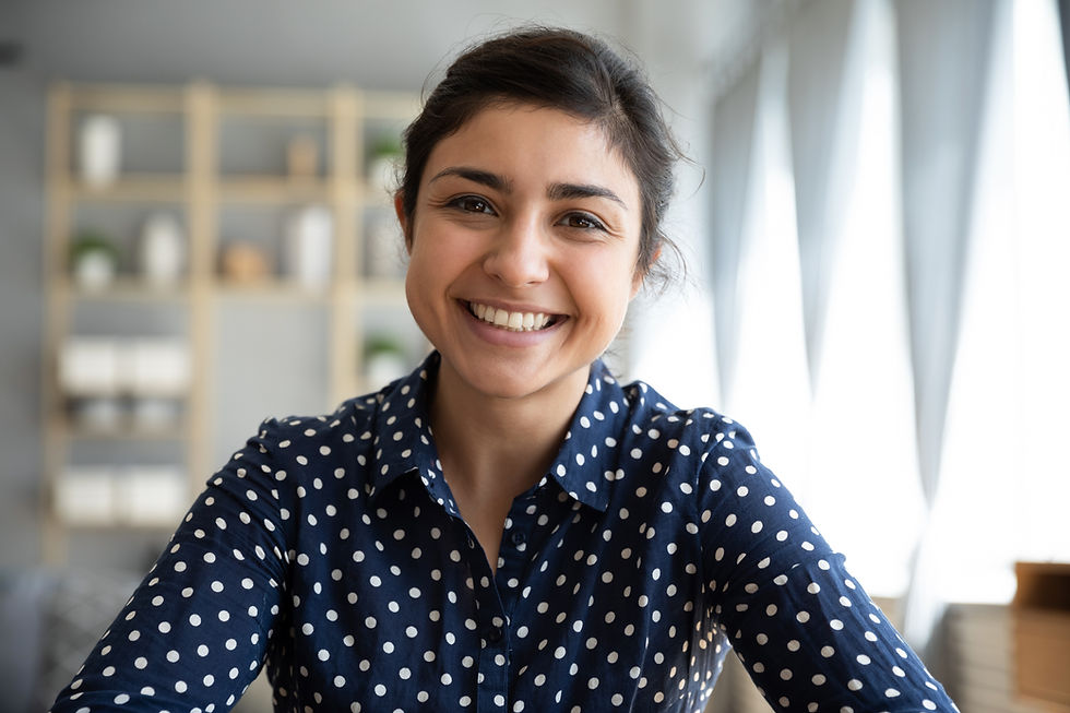 This is a picture of a smiling woman. She has black hair pulled away from her face, she has olive-colored skin, and she's wearing a navy blue top with white polka dots. In the background is a gray wall with a shelf full of objects.