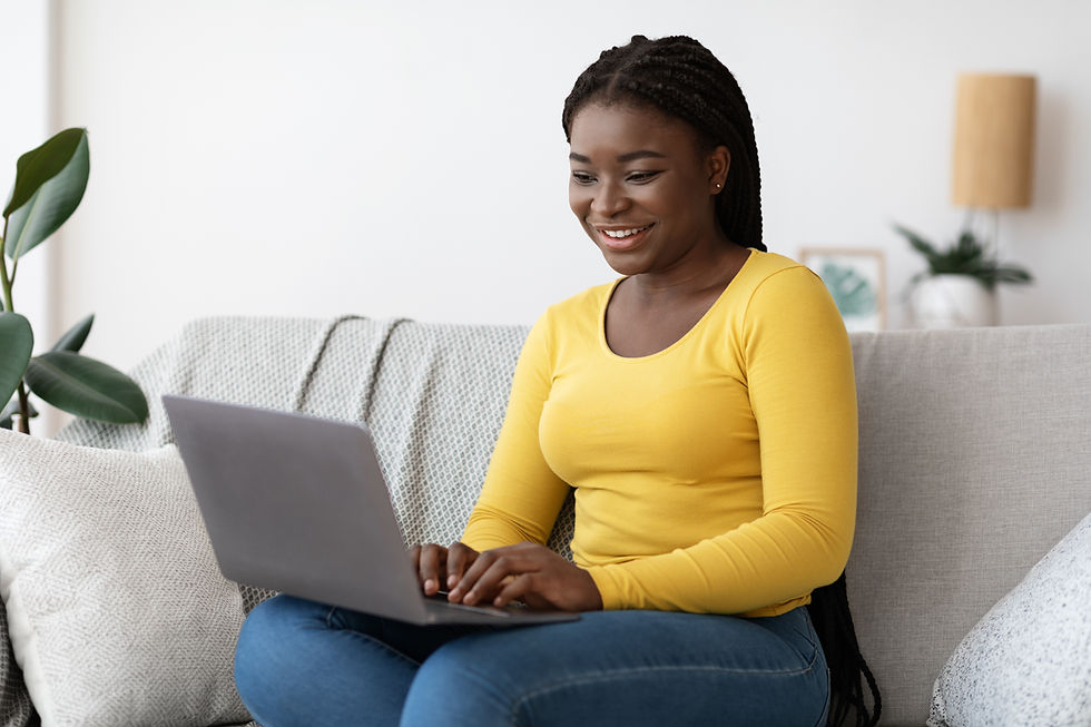 This is a picture of a Black woman sitting on a gray couch. She's wearing jeans and a long-sleeved yellow shirt. On her lap is an open laptop.