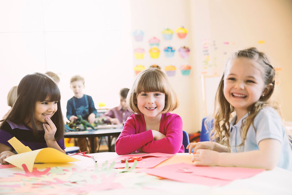 Three girls are sitting at a table in a classroom. There are craft supplies strewn about on the table, and all three girls are looking at the camera.