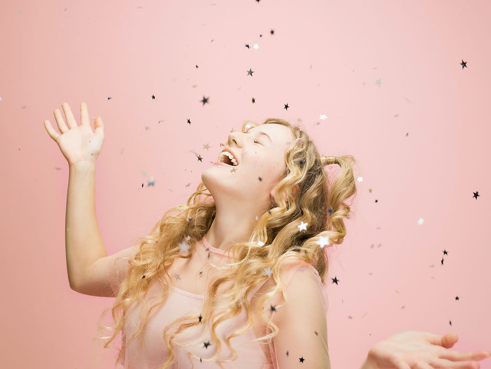 A young teenage girl with blond curly hair is wearing a light pink sleeveless dress against a matching background. She has thrown silver, star-shaped confetti into the air, and she is smiling. 