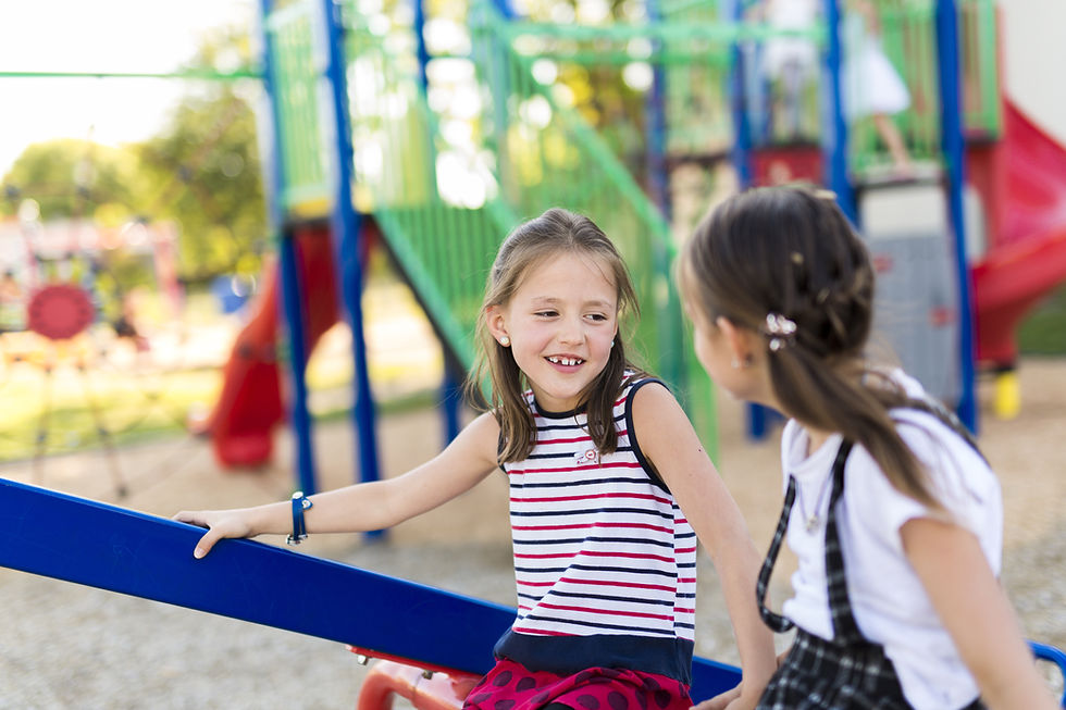 This is a picture of two caucasian girls sitting together by a playground. Both girls have long, straight brown hair, and they are looking at each other. The girl on the left is wearing a red, white and blue striped tank top, as well as a red skirt with navy blue polka dots. Her face can be seen. The girl on the right is wearing a white, short-sleeved shirt with a plaid pinafore. In the background is a jungle gym that is colored blue, green, red, black, and gray.