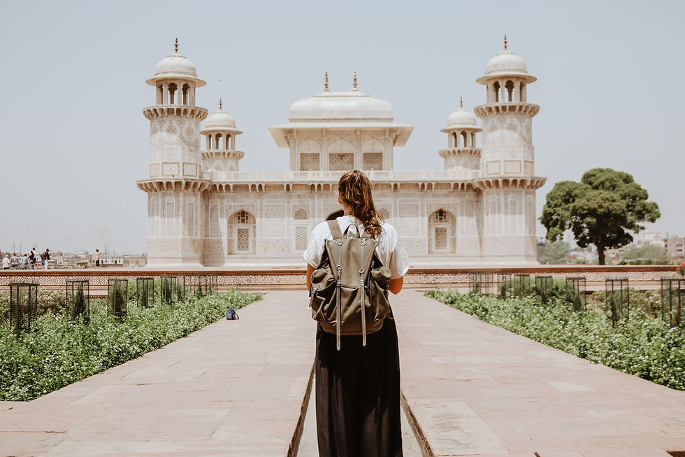 This is a picture of a woman standing in front of an ancient Indian building. Her back is towards the camera, she's wearing a backpack, and her hair is in a ponytail.