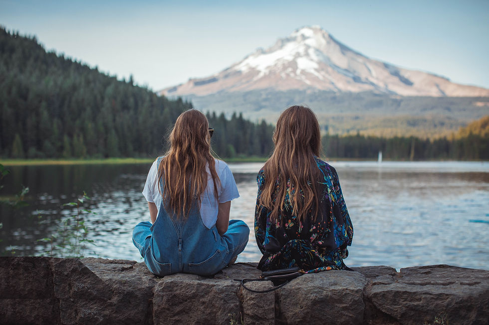 This is a picture of two young women sitting on a rock wall with their backs to the camera. In the distance is a lake, a forest, and a mountain. 