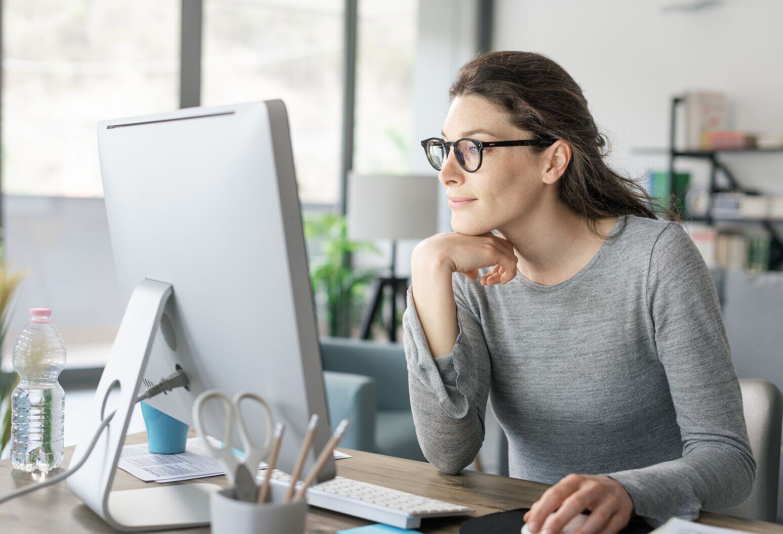 Woman with dark hair, glasses, and a gray shirt at a computer monitor.