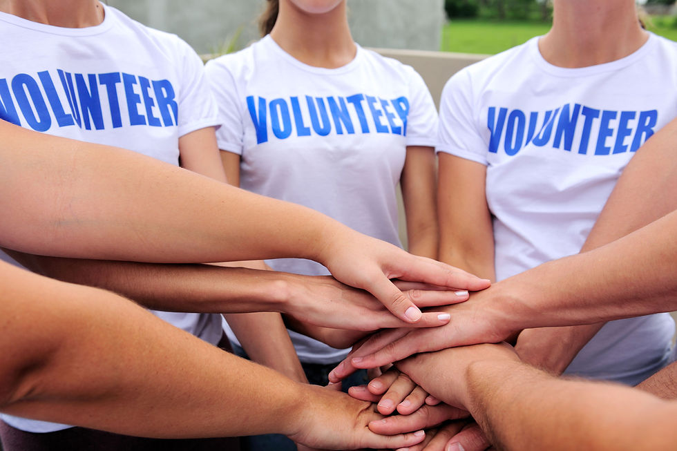 There are three people in the picture with two people off-camera. The people in the picture are wearing white shirts with blue block letters that say "Volunteer." Everyones arms are outstretched in front of them, and their hands are stacked on top of each other.
