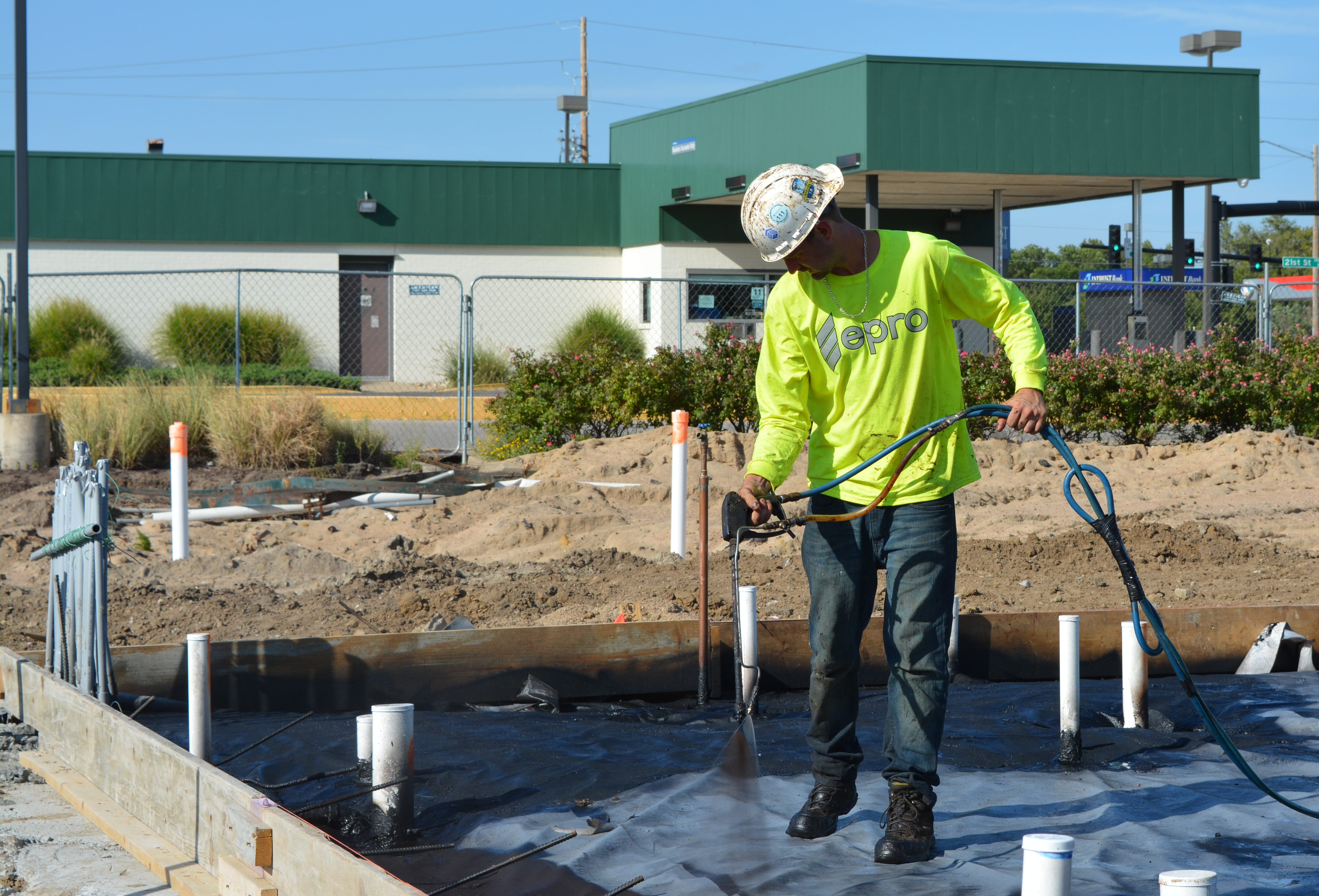 A construction site with a person in a yellow shirt and white hard hat spraying EPRO Geo-Seal Bond on a vapor intrusion barrier system.
