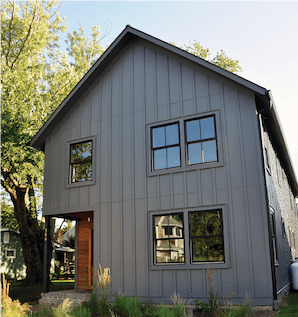 A house with vertical grey siding and wood posts in the front with black windows and a forest backgr…